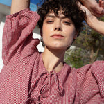 Woman wearing a red and white checkered linen dress with arms raised outdoors.