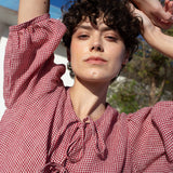 Woman wearing a red and white checkered linen dress with arms raised outdoors.