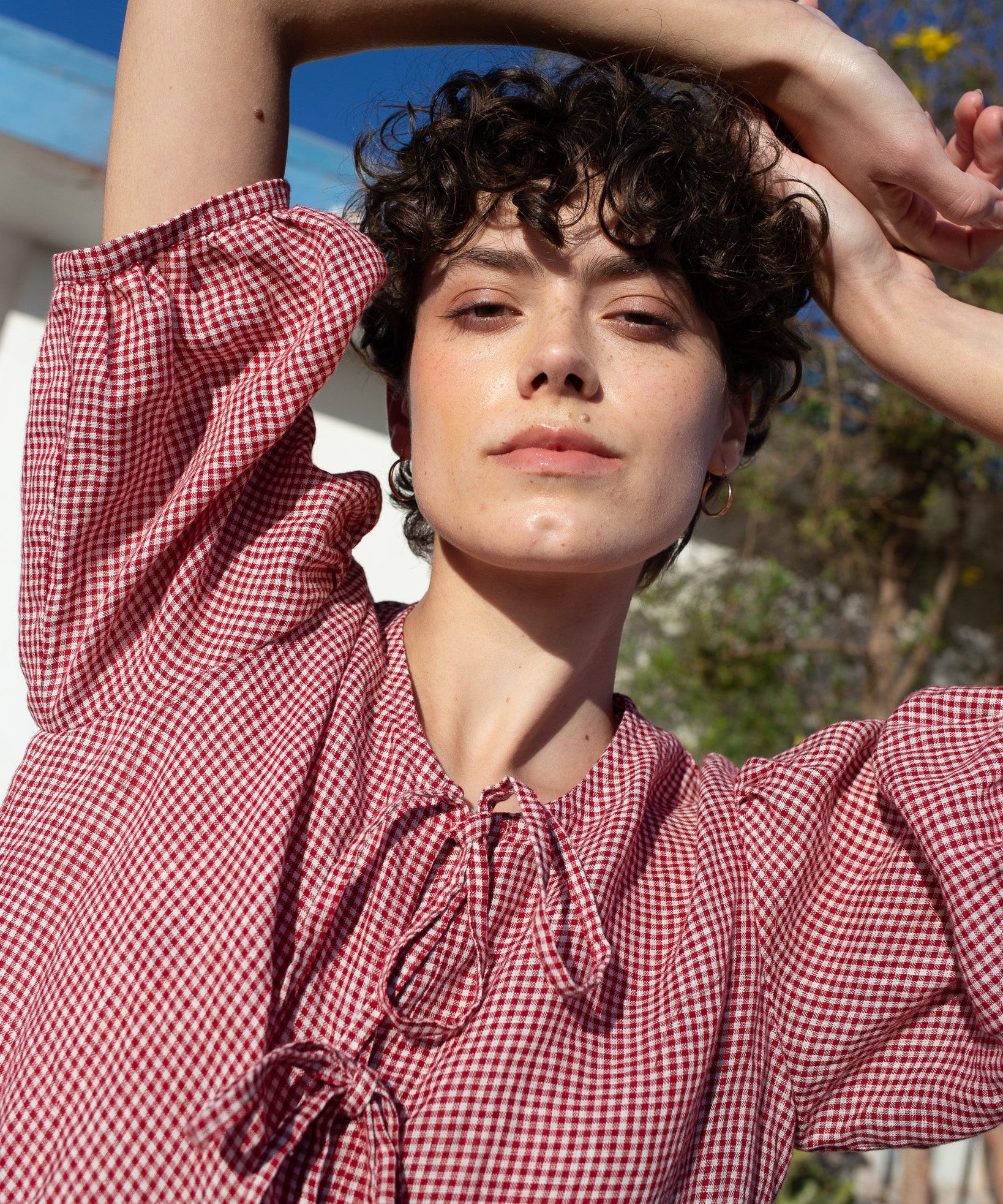 Woman wearing a red and white checkered linen dress with arms raised outdoors.