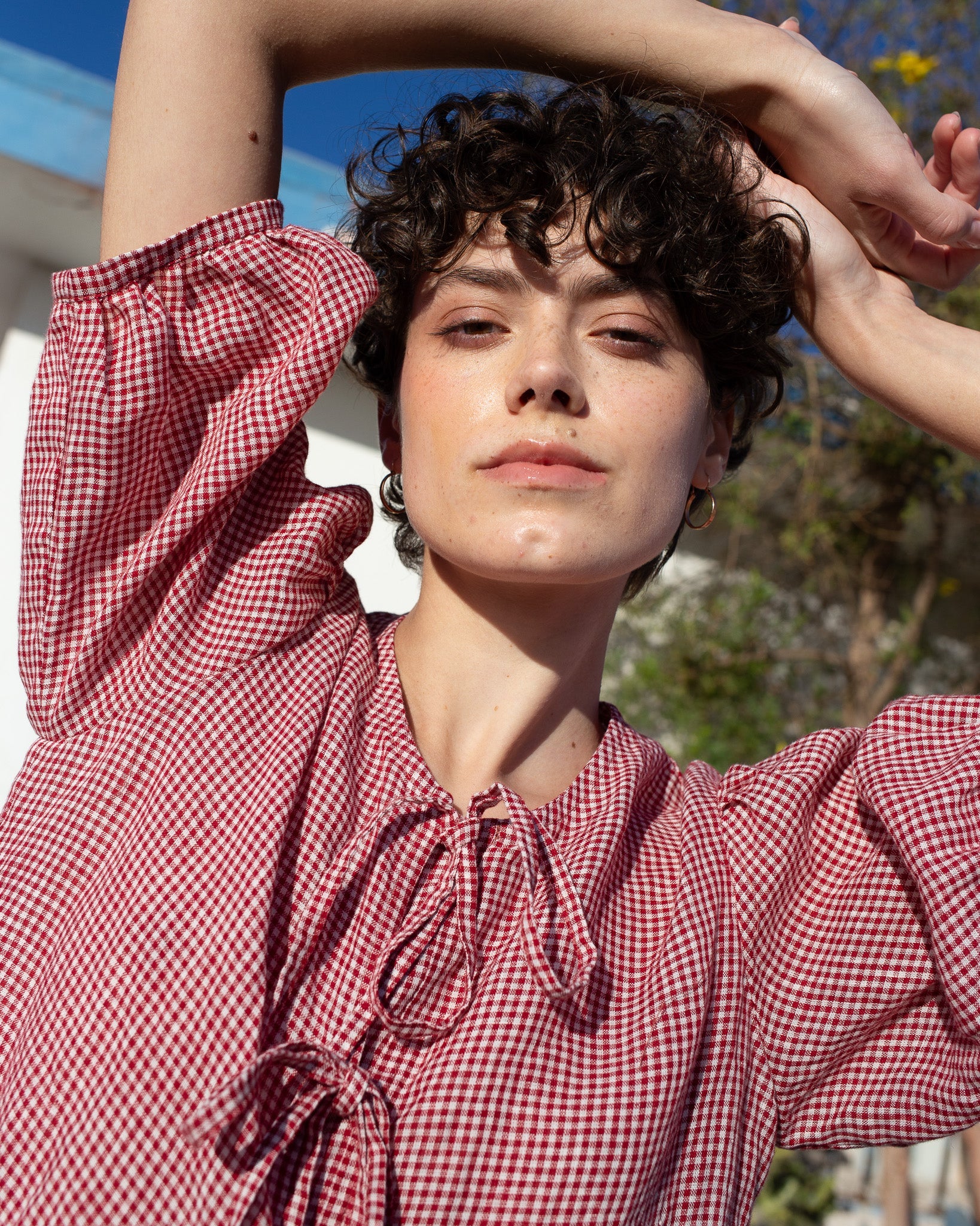 Woman wearing a red and white checkered linen dress with arms raised outdoors.